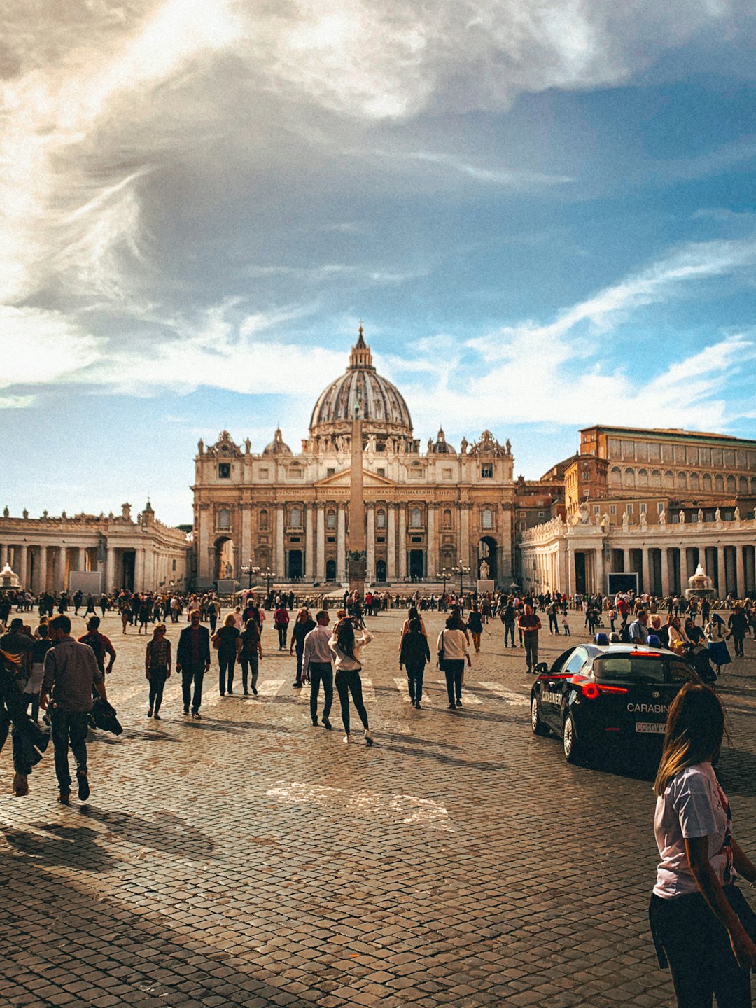 Folla eterogenea attraversa una piazza acciottolata con una basilica sullo sfondo.